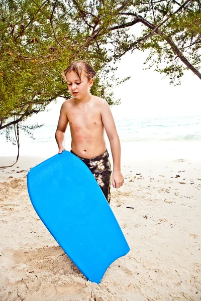 Мальчик на пляже с доской для серфинга улыбается и смотрит — стоковое фото