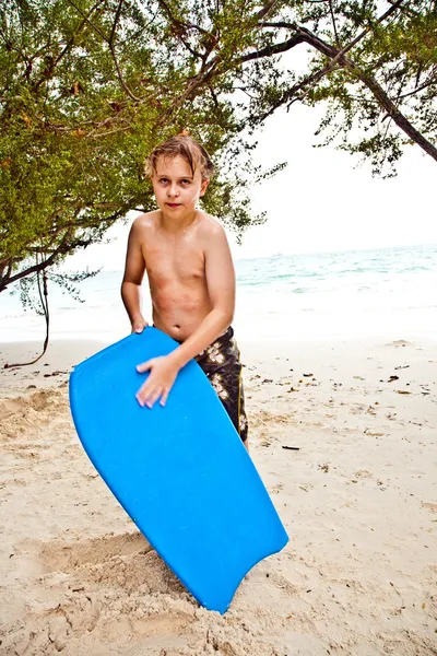 Niño en la playa con tabla de surf está sonriendo y mirando se —  Fotos de Stock