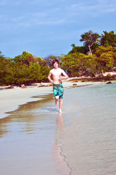 Młody chłopak z czerwone włosy w strój kąpielowy jest biegnącej wzdłuż pięknej plaży — Zdjęcie stockowe