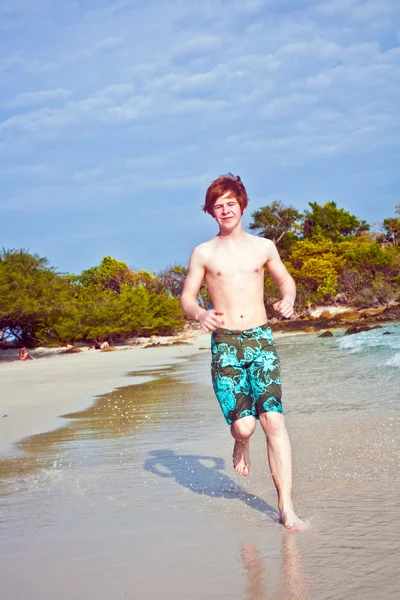 Ung pojke med rött hår i baddräkt kör längs den vackra stranden — Stockfoto