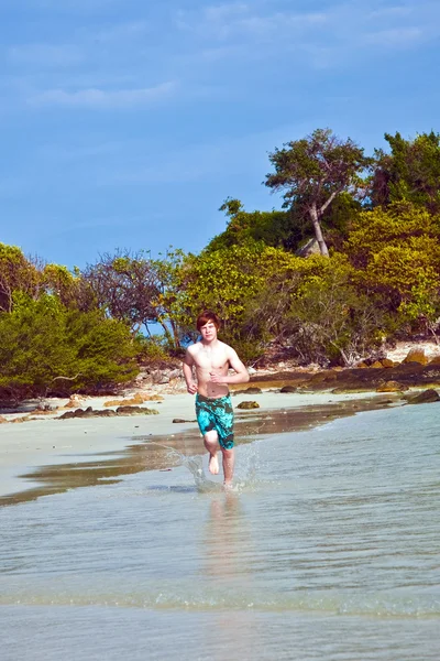 Młody chłopak z czerwone włosy w strój kąpielowy jest biegnącej wzdłuż pięknej plaży — Zdjęcie stockowe