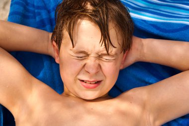 Cute young boy at the beach closes eyes because of bright sunlig clipart