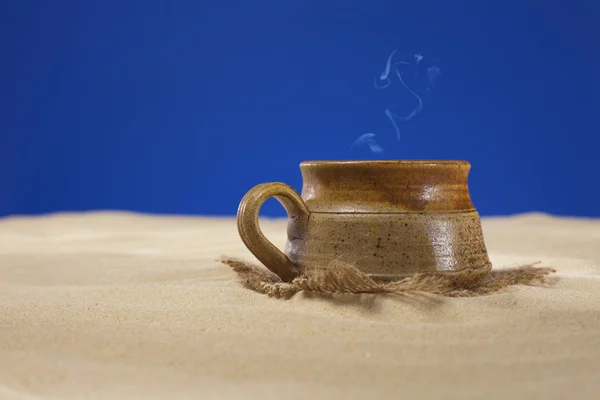 Clay mug with tea or coffee on beach sand, blue background — Stock Photo, Image