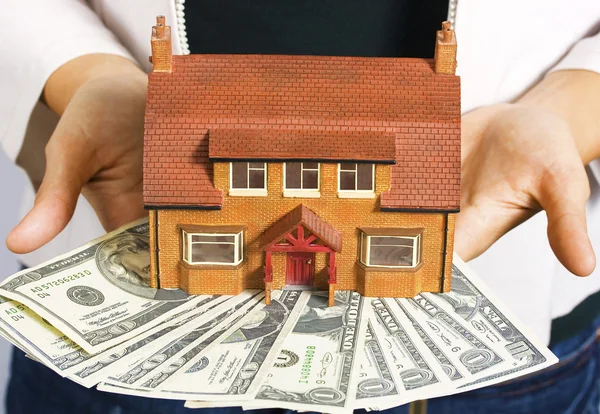 Uma pessoa segurando uma casa em miniatura e algumas notas de dólar — Fotografia de Stock