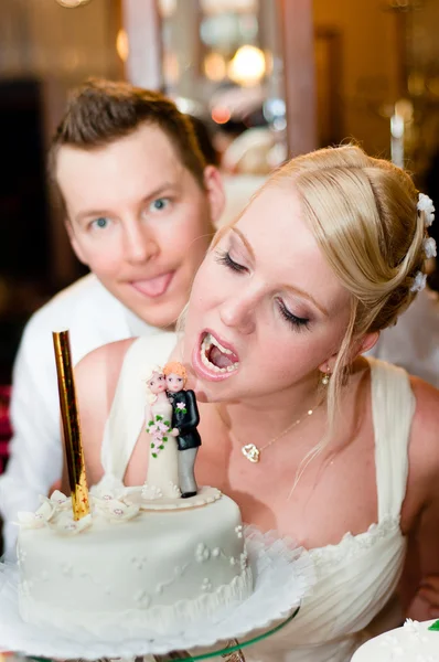 Junge Braut wird mit Bräutigam im Hintergrund in ihren Kuchen beißen — Stockfoto