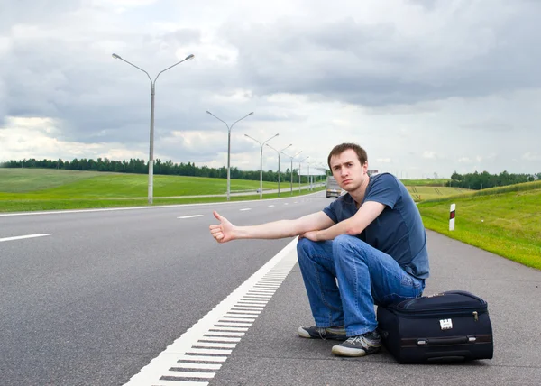 stock image The young man sits pending on road with a suitcase