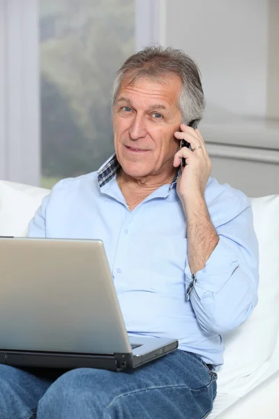 Hombre mayor en sofá con ordenador portátil y teléfono móvil — Foto de Stock