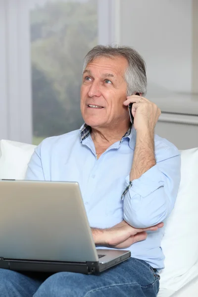 Hombre mayor en sofá con ordenador portátil y teléfono móvil — Foto de Stock