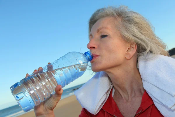 Aktivní senior žena pitnou vodu z láhve na pláži — Stock fotografie
