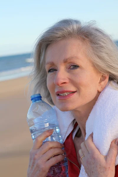 Mulher idosa ativa bebendo água da garrafa na praia — Fotografia de Stock