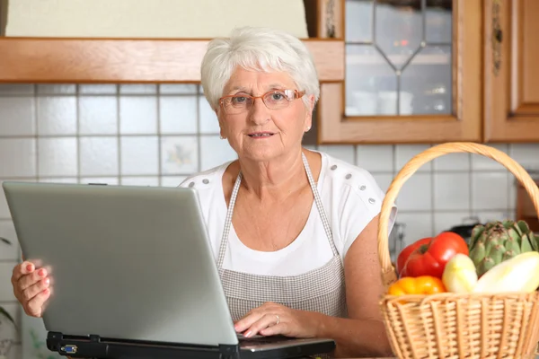 Femme âgée dans la cuisine avec ordinateur portable — Photo