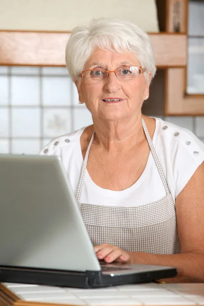 Äldre kvinna i köket med bärbar dator — Stockfoto
