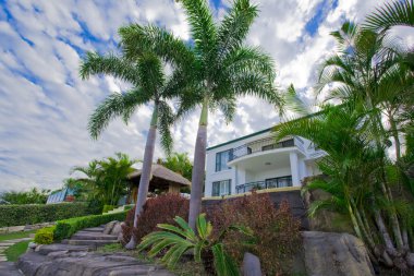 Garden with palms and Bali hut in front of waterfront mansion clipart