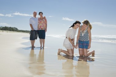 Sisters looking at shell on beach with parents in background clipart