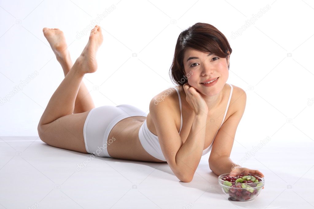 Full body shot of beautiful, young oriental girl, lying on floor wearing white sports underwear. Model has fit healthy body and is holding a bowl of fruit.