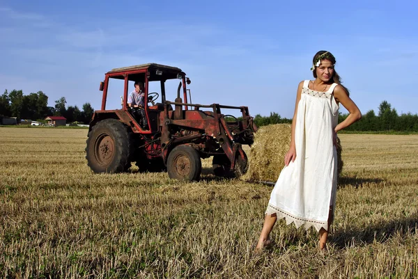 Dívka v venkovské oblečení stojí na poli, traktor na pozadí — Stock fotografie