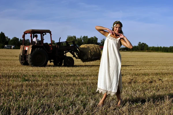 Κορίτσι σε μια αγροτική ενδυμασία στέκεται στο πεδίο, τρακτέρ στο παρασκήνιο — Φωτογραφία Αρχείου