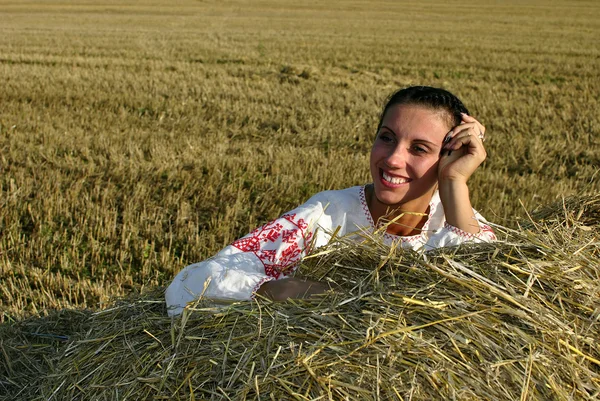 Κοπέλα με παραδοσιακή ρωσική φορεσιά που βρίσκεται σε μια θυμωνιά χόρτου και χαμογελαστός — Φωτογραφία Αρχείου