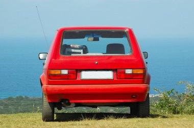 Arkadan görünüşü ayakta açık havada mavi gökyüzü önünde çimenlikte park etmiş bir küçük kırmızı araba ve (Vw Citi Golf Servisi)