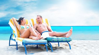 Rear view of a couple on a deck chair relaxing on the beach clipart