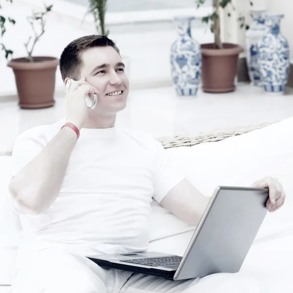 Uśmiechający się młody człowiek pracy na komputerze przenośnym i połączenia przez telefon — Zdjęcie stockowe