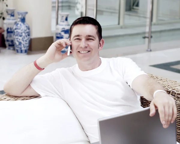 Lächelnder junger Mann, der am Laptop arbeitet und telefonisch einen — Stockfoto