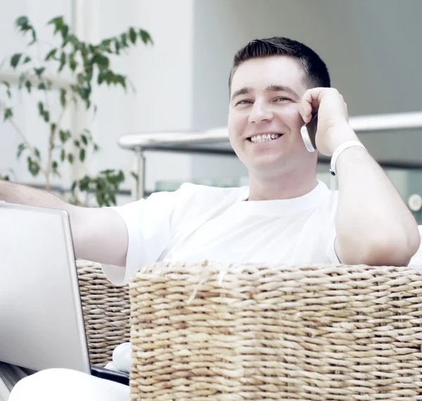 Улыбающийся молодой человек, работающий на ноутбуке и звонящий по телефону — стоковое фото