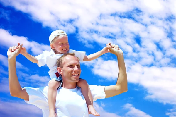 Vrolijke vader piggyback rit geven aan zijn zoon tegen een hemel bac — Stockfoto