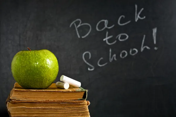 Chalk and green apple on old textbook against blackboard — Stock Photo, Image