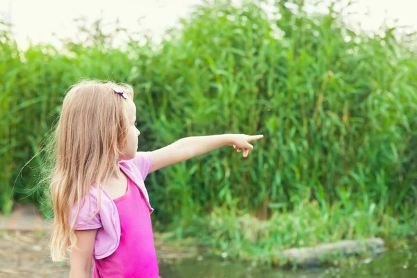 Blond dziewczynka pokazuje Naciągnij je na bok, w pobliżu rzeki z laski — Zdjęcie stockowe
