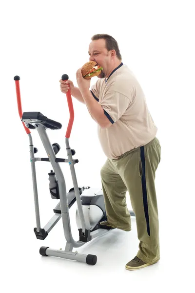 Overweight man eating by an exercising device — Stock Photo, Image