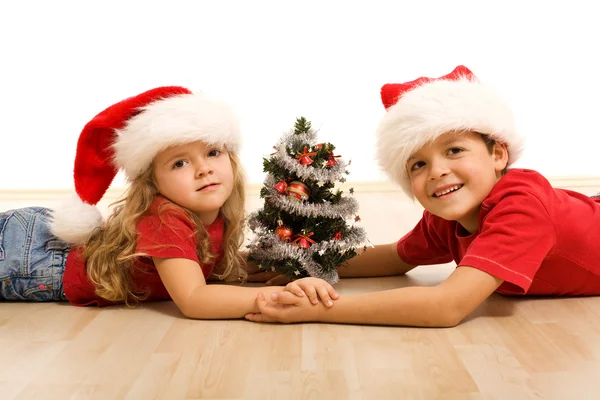 Niños en el suelo con un árbol decorado y sombreros de Navidad —  Fotos de Stock