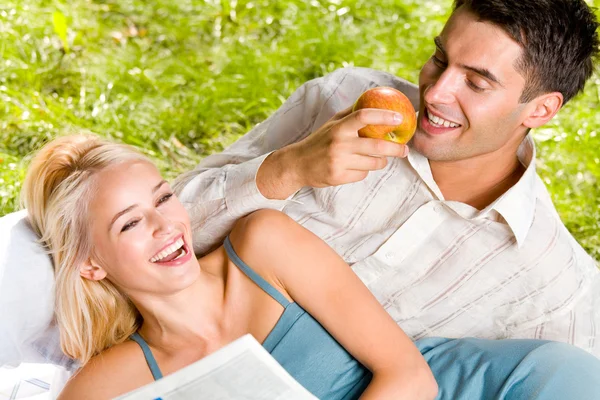 Joven pareja feliz comiendo manzanas y leyendo el periódico al aire libre — Foto de Stock