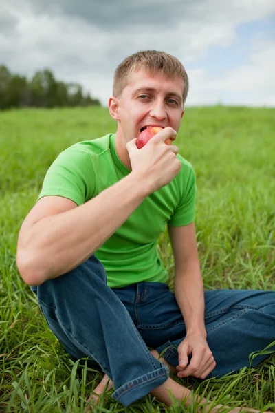 Young man sitting on the grass and biting apple — Stock Photo, Image