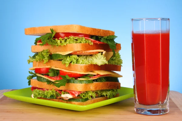Grande sanduíche e vidro de suco de tomate no fundo azul — Fotografia de Stock
